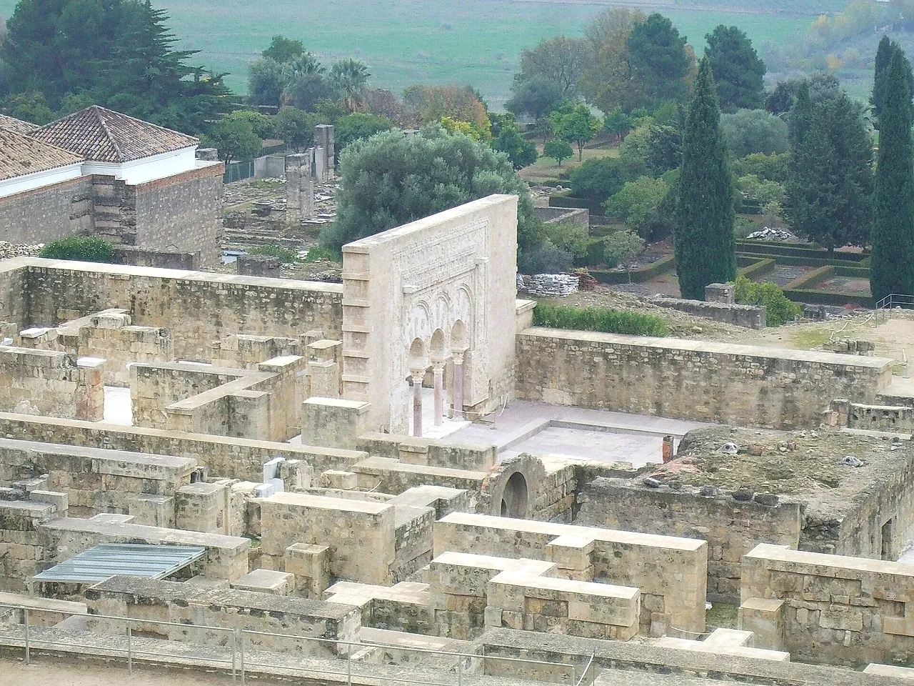 Overview of the excavated ruins of Medina Azahara, with rows of stone walls, a decorated façade and cypress trees beyond