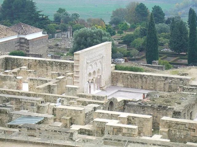 Overview of the excavated ruins of Medina Azahara, with rows of stone walls, a decorated façade and cypress trees beyond