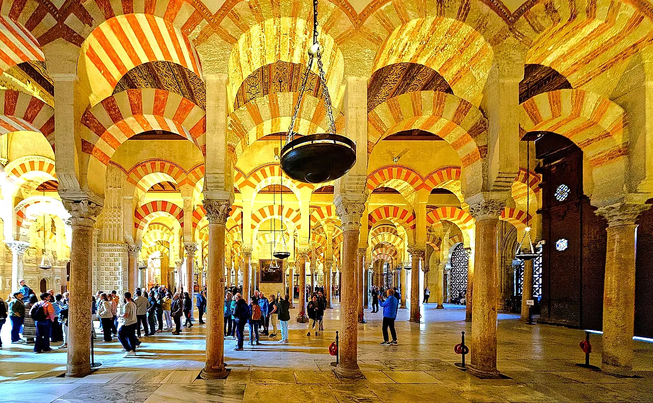 Rows of columns and two-tier red-and-white arches inside the Mosque-Cathedral of Córdoba, with a hanging lamp in the centre and visitors walking between the columns