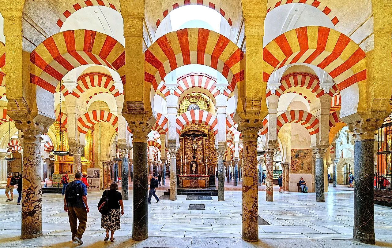A long aisle of red-and-white arches inside the Mosque-Cathedral of Córdoba, with a lit chapel at the far end and visitors in the foreground